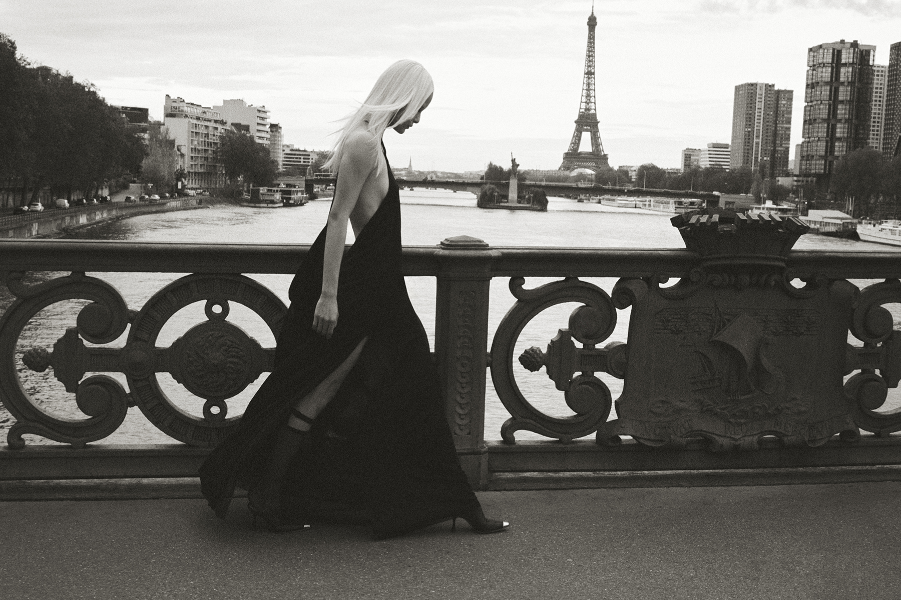 Black and white fashion photograph of a model in a long black dress walking across a Paris bridge with the Eiffel Tower in the background